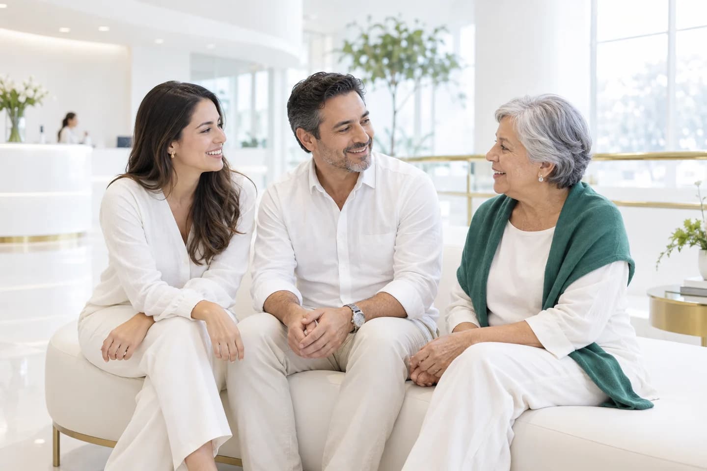 Personas de distintas generaciones conversan en una recepción luminosa de salud, blanca, con detalles verdes y dorados